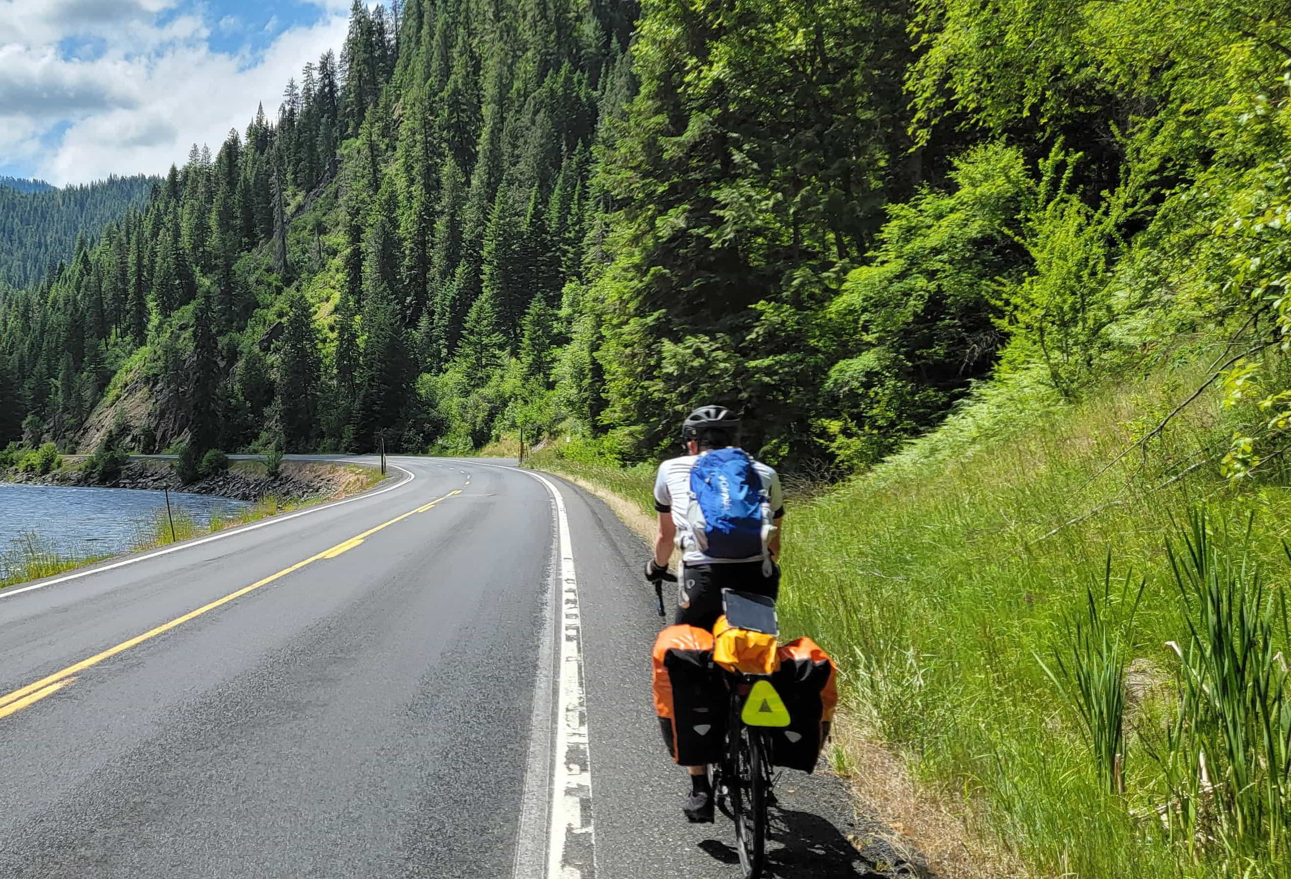 A cyclist with panniers rides on a scenic road with trees to his right, and a winding river to his left